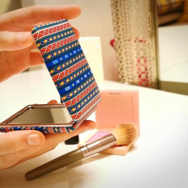 Person holding a foldable compact mirror with vibrant tribal pattern cover, placed on a makeup vanity with blush and brush.