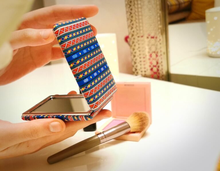Person holding a foldable compact mirror with vibrant tribal pattern cover, placed on a makeup vanity with blush and brush.