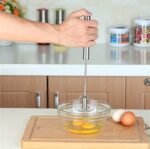 "Hand using stainless steel push whisk to mix eggs in a glass bowl – self-rotating manual egg beater with ergonomic grip"