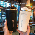 "Two stainless steel insulated coffee travel mugs in black and white with flip-top lids, held by hand in a modern café setting"